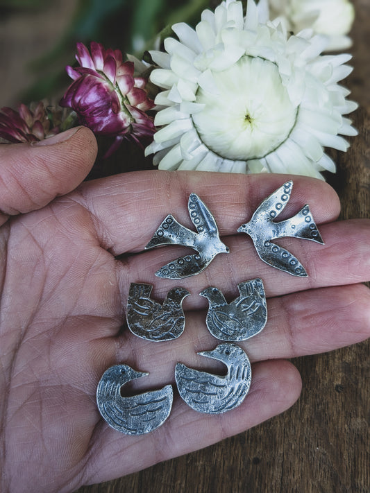 Hand holds three sterling silver earring pairs featuring flying birds, loons and chickens stamped in a folk art style