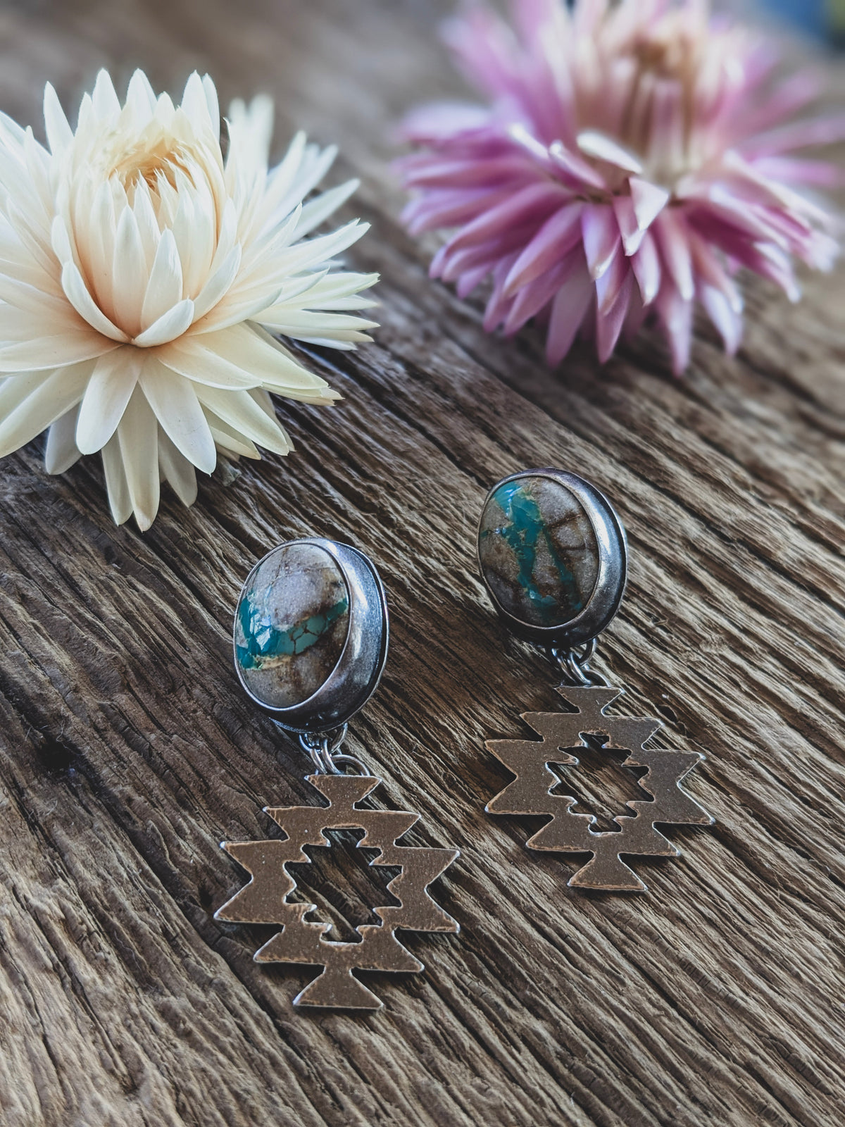 A pair of stud earrings on a board, in front of dried flowers. The stones are white with turquoise colored seams running through them and the earrings have a geometric dangle component. 