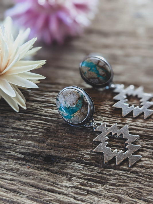 A pair of stud earrings on a board, in front of dried flowers. The stones are white with turquoise colored seams running through them and the earrings have a geometric dangle component. 