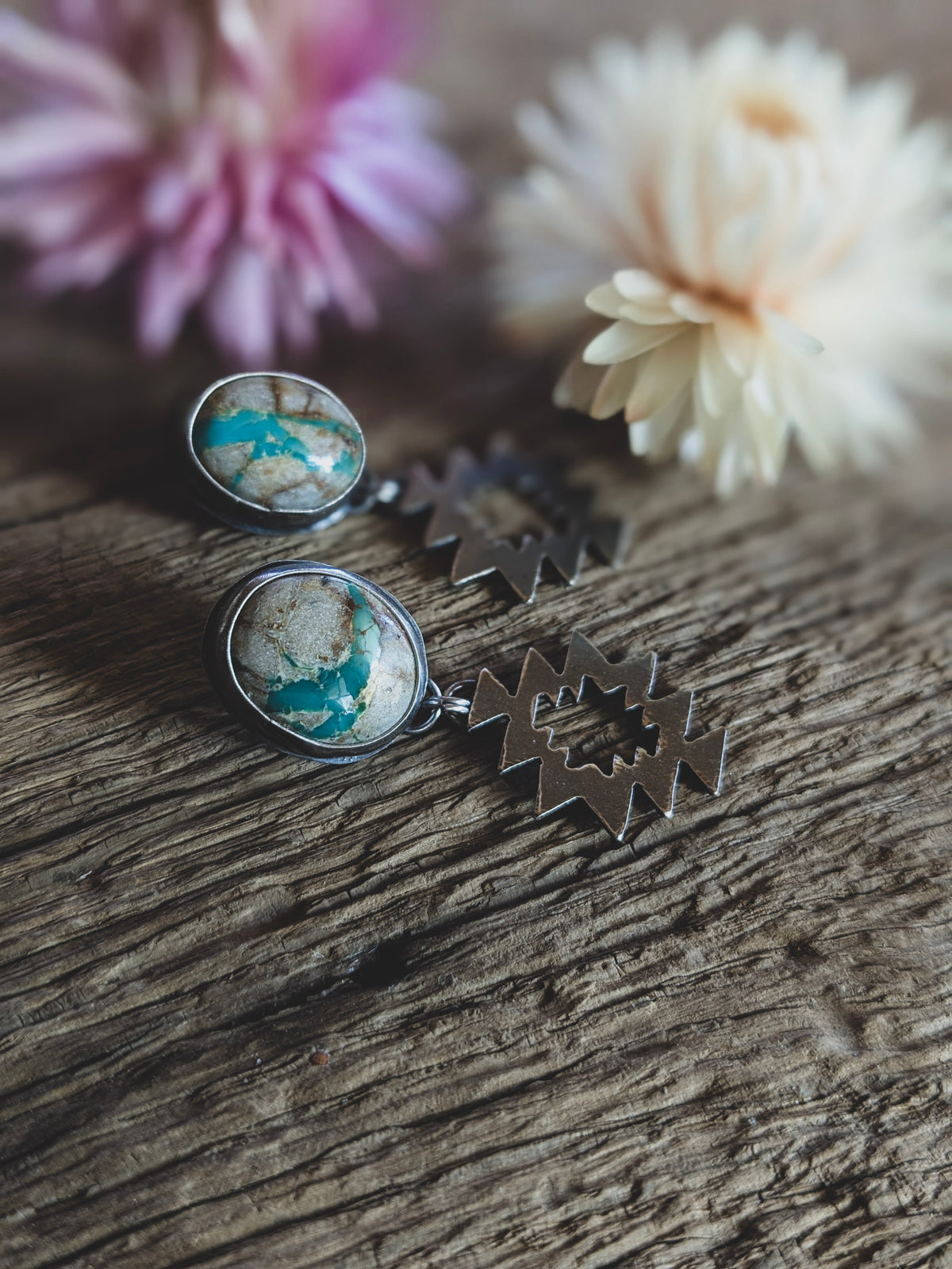 A pair of stud earrings on a board, in front of dried flowers. The stones are white with turquoise colored seams running through them and the earrings have a geometric dangle component. 