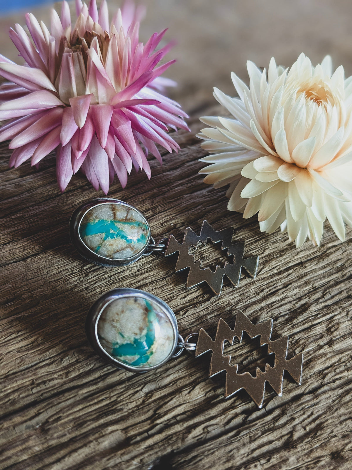 A pair of stud earrings on a board, in front of dried flowers. The stones are white with turquoise colored seams running through them and the earrings have a geometric dangle component. 